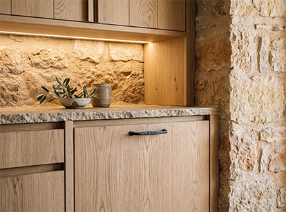 Kitchen detail: oak fronts, a granite worktop and a limestone wall lit by grazing light.