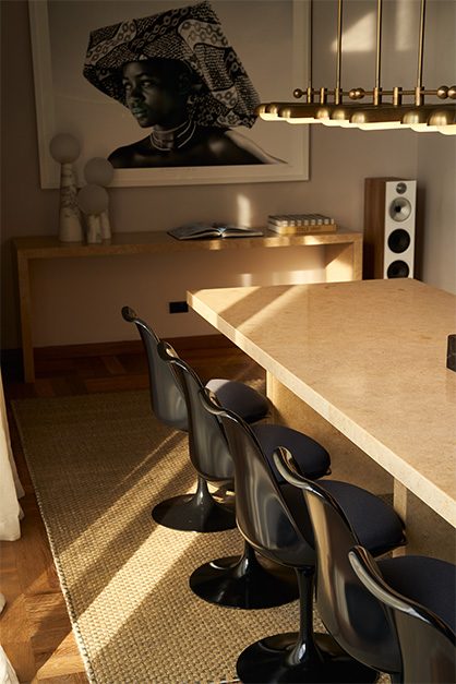 A dining room with a pale stone table, black designer chairs and a framed portrait beneath a linear brass pendant.