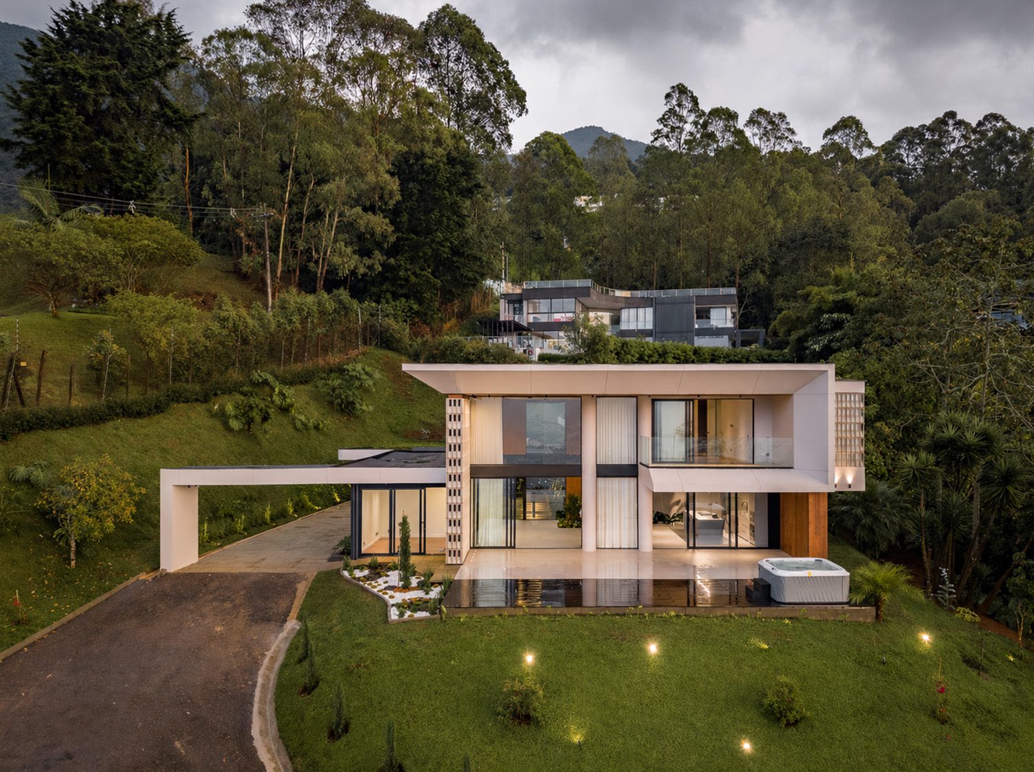 Casa Asturias at dusk: a two-storey volume with a glazed façade, a sloping garden and a swimming pool, surrounded by woodland.